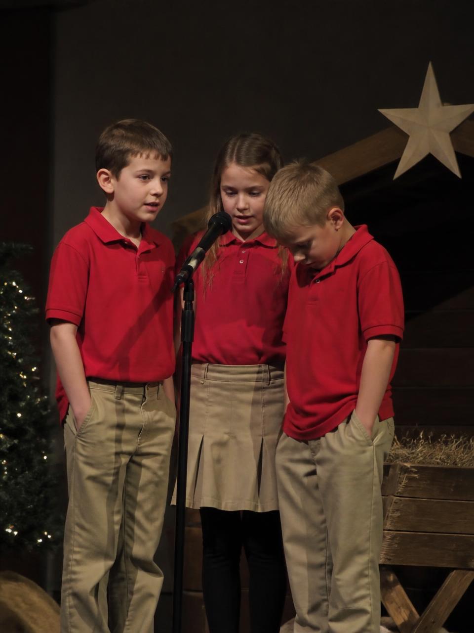 Illustration of three students singing at a microphone beside the manger set during the Christmas program